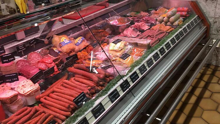 Eine Metzgereitheke mit verschiedenen Fleisch- und Wurstsorten, beschriftet mit Preisschildern. Die Auslage wirkt gut gefüllt und vielfarbig arrangiert.