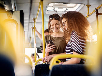 Two cheerful pretty young women are standing in a bus and looking at the phone and smiling while waiting for a bus to take them to their destination.