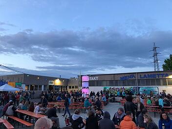 Das Bild zeigt eine entspannte Outdoor-Veranstaltung mit bunten Lichtern und Graffiti. Menschen sitzen an Tischen und genie&szlig;en die Atmosph&auml;re eines Festivalabends.
