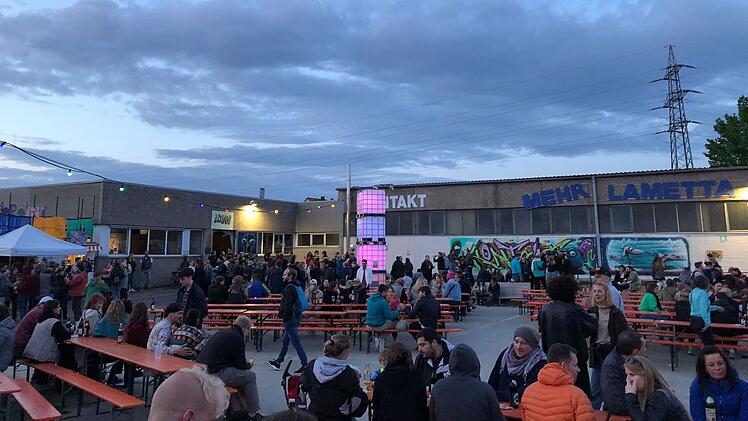 Das Bild zeigt eine entspannte Outdoor-Veranstaltung mit bunten Lichtern und Graffiti. Menschen sitzen an Tischen und genießen die Atmosphäre eines Festivalabends.