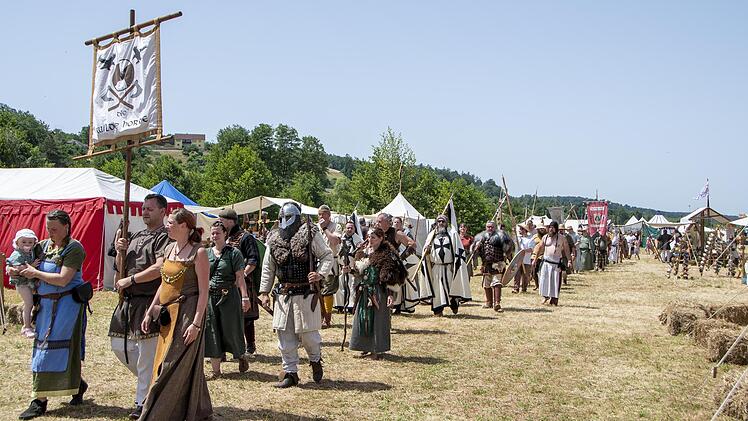 Eine Gruppe von Menschen in mittelalterlichen Gewandungen marschiert über ein Feld, umgeben von Zelten, mit einem Banner vorneweg. Sie nehmen an einem historischen Lager teil.