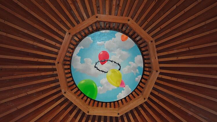 Die Kirchendecke scheint sich zu öffnen. Ein Stück blauer Himmel mit weißen Wolken wird sichtbar. Luftballons steigen hinauf in farbenfroher Leichtbarkeit. Werden sie zerplatzen an der Dornenkrone? Wie das Kreuz im Altarraum vergegenwärtigen sie Jesu Leiden und Sterben. (aus der Begleitbroschüre)