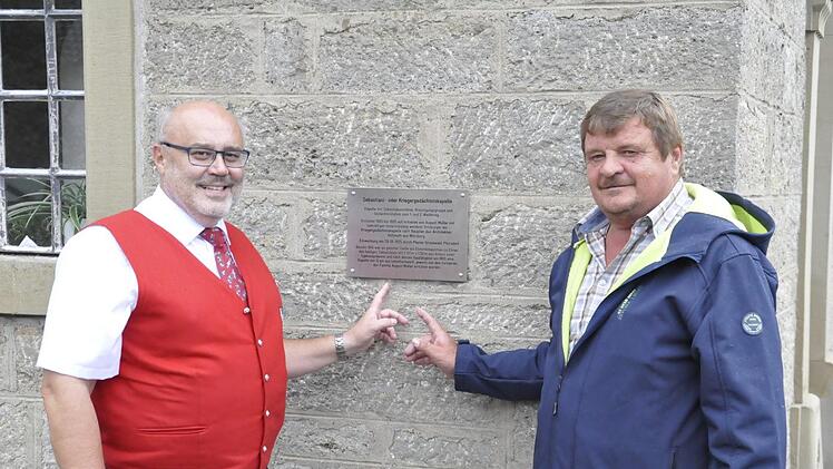 Bürgermeister Franz Kuhn (links) und der Vorsitzende der Oerlenbacher Jagdgenossenschaft Ekkehard Schmitt (rechts) enthüllen die Gedenktafel zur Geschichte der Sebastiani- oder Kriegergedächtniskapelle.