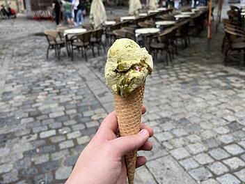 Eine Hand hält ein Pistazieneis in einer Waffel auf einem gepflasterten Platz mit mehreren Tischen und Stühlen im Hintergrund.