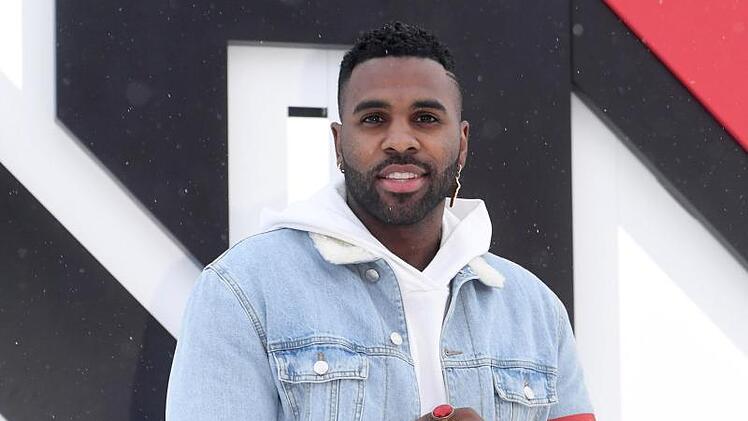 Ein Mann in einer Jeansjacke, wei&szlig;em Hoodie und mit viel Schmuck posiert l&auml;chelnd vor einer grafischen Kulisse. Es k&ouml;nnte sich um den angek&uuml;ndigten Weltstar f&uuml;r das Event handeln.