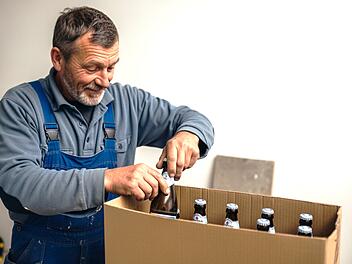 Ein Mann in blauer Arbeitskleidung packt lächelnd Bierflaschen in einen Karton. Es scheint, als würde er fränkisches Bier für den Versand vorbereiten.