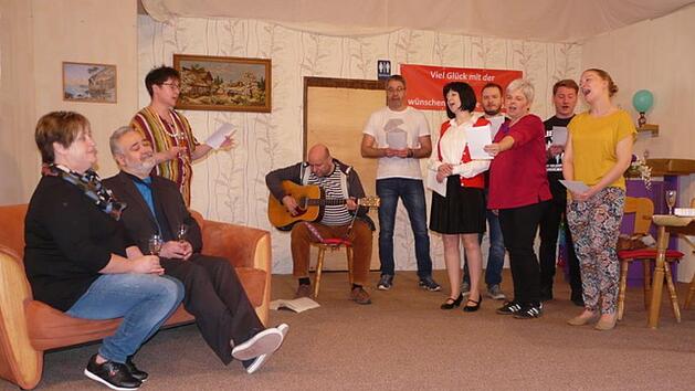 Mit einem Lied verabschieden die Arbeitskollegen Julius Klein, der mit seiner Frau Therese auf dem Sofa sitzt, in den Ruhestand. Seine ehemalige Sekret&auml;rin Lieselotte Schl&uuml;pfer dirigiert den Chor. Foto: Karin G&uuml;nther