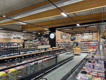 Moderner Supermarkt mit Holzakzenten und vielen Produkten. Kühltruhen und Regale gut gefüllt. Beleuchtung und große Gänge erleichtern das Einkaufen. Einladende Atmosphäre.