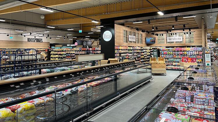 Moderner Supermarkt mit Holzakzenten und vielen Produkten. Kühltruhen und Regale gut gefüllt. Beleuchtung und große Gänge erleichtern das Einkaufen. Einladende Atmosphäre.
