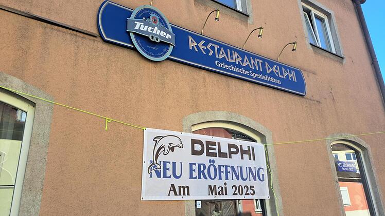 Ein griechisches Restaurant namens "Delphi" kündigt seine Neueröffnung im Mai 2025 an. Ein blaues Schild mit "Restaurant Delphi" ist sichtbar, darunter ein Banner mit der Ankündigung.