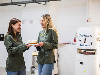 Zwei Frauen in gr&uuml;nen Jacken halten eine Torte und lachen in einer B&auml;ckerei. Im Hintergrund steht eine wei&szlig;e Maschine mit blauer Aufschrift.